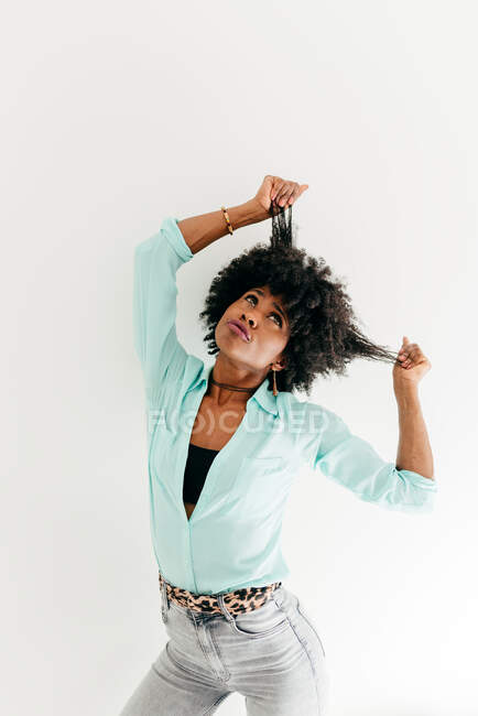 Playful young African American female in trendy outfit having fun touching afro hair looking up on white background — Stock Photo
