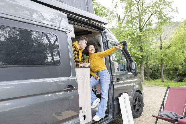 Happy black woman with beer smiling and pointing away while standing in van with boyfriend during road trip in countryside — Stock Photo