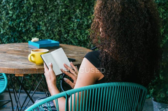 Back view of unrecognizable curly haired female entrepreneur sitting at table with cup of coffee and smartphone and flipping empty pages of planner on cafe terrace — Stock Photo