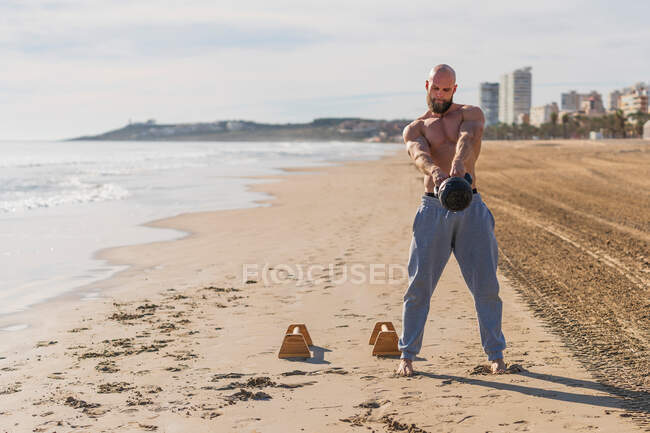 Full body bald and shirtless man doing swings with kettlebell while standing barefoot on sandy coast with city on background — Stock Photo