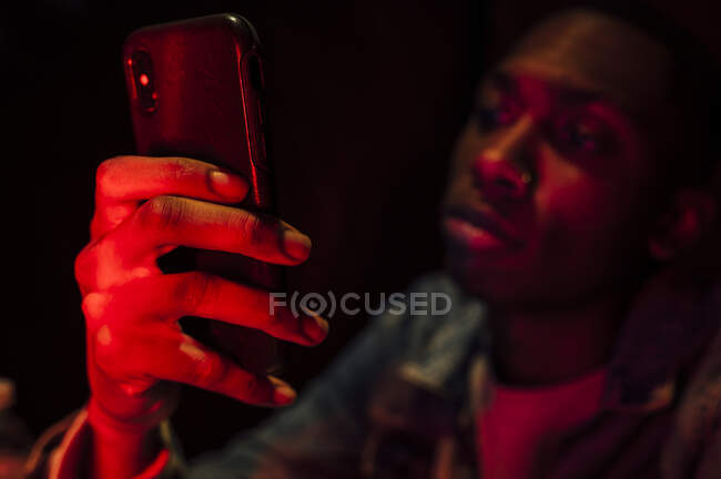 Side view of crop African American calm serious guy in denim jacket calling on mobile phone in neon mysterious red light on black background — Stock Photo