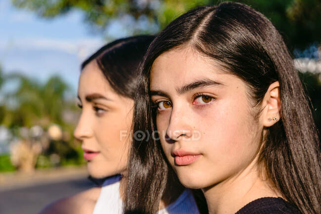 Adolescente con i capelli castani e gli occhi vicino sorella femmina in giornata di sole su sfondo sfocato — Foto stock