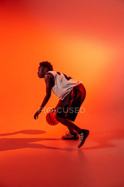 Mujer negra con traje de baloncesto en el estudio usando geles de color y luces del proyector sobre fondo naranja - foto de stock