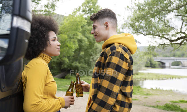 Vista lateral do jovem multirracial e mulher batendo garrafas de cerveja e olhando uns para os outros enquanto relaxa na natureza no dia de verão — Fotografia de Stock