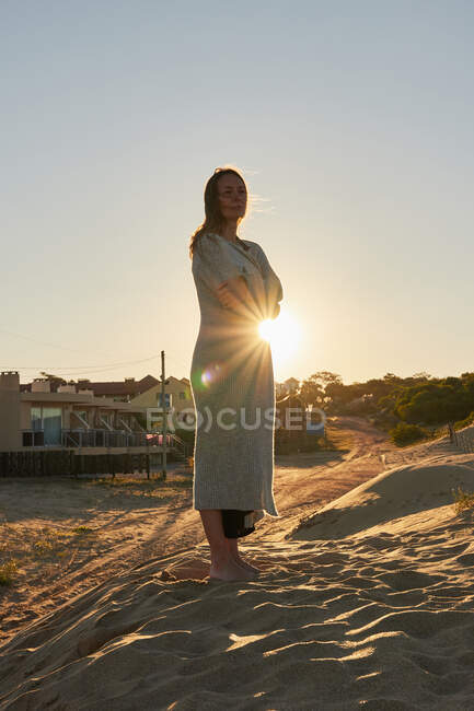 Mujer pacífica con los ojos cerrados de pie en la orilla arenosa en el fondo del brillante cielo del atardecer en verano - foto de stock