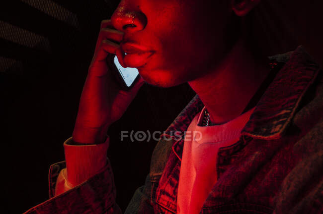Зворотний бік афроамериканців спокійний серйозний хлопець у джинсовій куртці, який дзвонить на мобільному телефоні з таємничим червоним світлом на чорному тлі. — стокове фото
