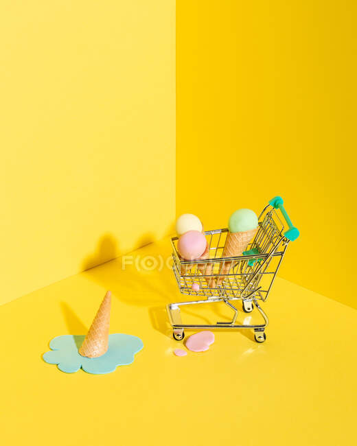 De composición anterior de carrito de compras en miniatura con una variedad de conos de helado multicolores colocados cerca de helado azul de fusión caído sobre fondo amarillo - foto de stock