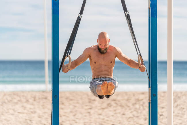 Мужчина без рубашки с бородой висит на гимнастических кольцах с поднятыми ногами, упорно тренируясь на песчаном пляже, глядя вниз — стоковое фото