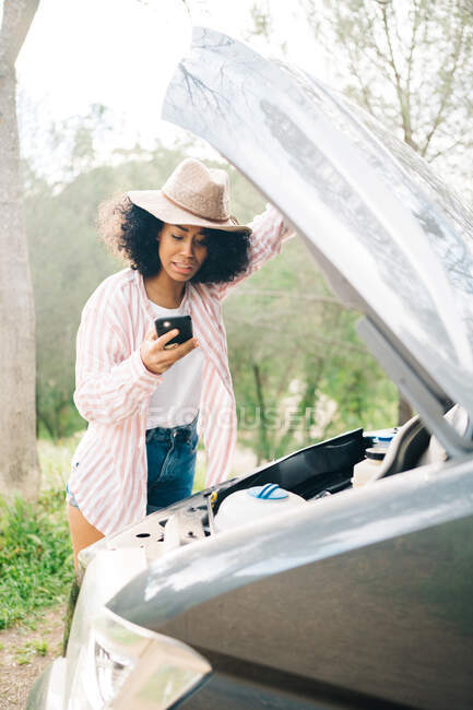 Молода афроамериканська жінка - мандрівниця, яка тримає смартфон з сяючим ліхтариком під час перевірки двигуна автомобіля як проблеми під час подорожі по природі. — стокове фото