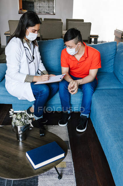 Весела жінка-практик в медичній сукні розмовляє з хлопчиком з синдромом Дауна і записує в буфері під час прийому вдома — стокове фото