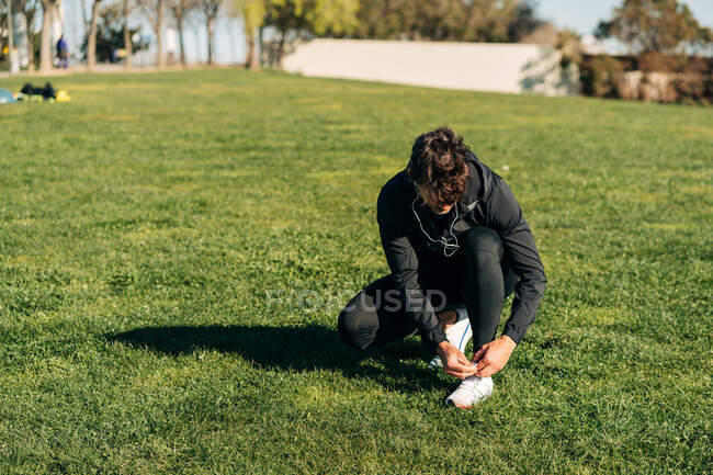 Atleta di sesso maschile anonimo in abiti sportivi che lega i lacci delle scarpe sulle calzature mentre si accovaccia sul prato prima dell'allenamento alla luce del sole — Foto stock
