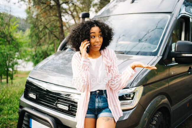 Sérieuse jeune femme afro-américaine parlant sur smartphone et gesticulant tout en s'appuyant sur le camping-car garé en forêt dans la journée d'été — Photo de stock