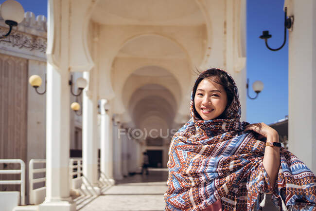 Joven hembra asiática positiva en colorido pañuelo tradicional sonriendo y mirando a la cámara mientras está de pie cerca del hermoso edificio blanco de la mezquita Al Rahma en Jeddah en Arabia Saudita - foto de stock