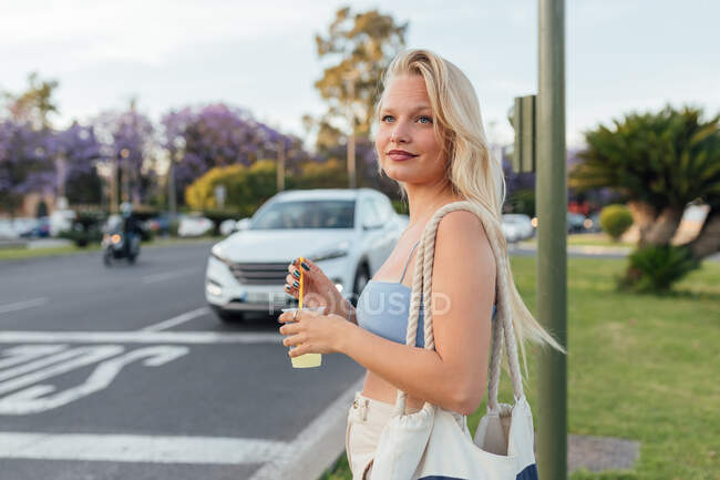 Side view of cheerful female standing with cold lemonade in plastic cup in street in summer — Stock Photo