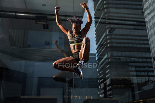 Side view of strong black woman jumping high near glass wall of modern building while exercising on city street on sunny day — Stock Photo