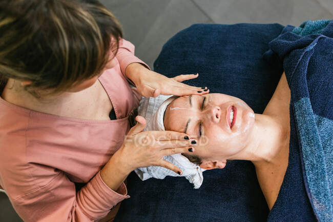 Da suddetto raccolto cosmetico irriconoscibile che applica il detergente facciale su faccia di cliente femminile durante trattamento di cura della pelle in salone di bellezza — Foto stock