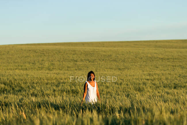 Smiling young black lady in white summer dress strolling on green wheat field while looking at camera in daytime under blue sky — Stock Photo
