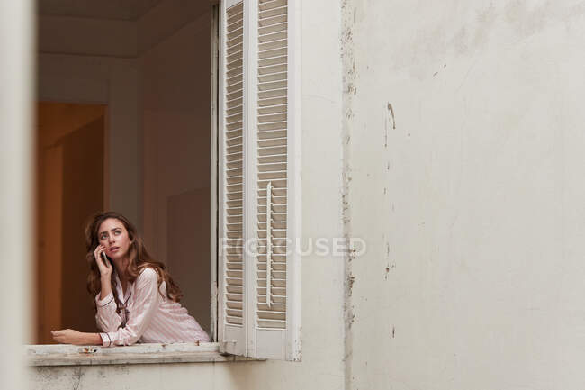 Mulher pacífica no pijama de pé perto da janela e falando no telefone celular em casa — Fotografia de Stock