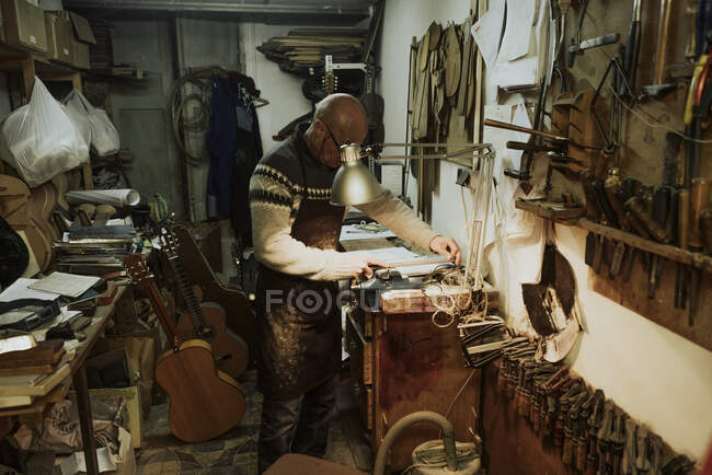 Vue latérale d'un artisan masculin chauve méconnaissable réparant des instruments à l'établi dans un petit studio d'art près de guitares acoustiques et de divers outils — Photo de stock