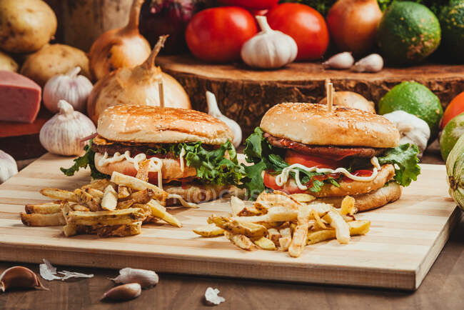 Hambúrgueres apetitosos com legumes e costeletas colocados em tábua de madeira com batatas fritas na cozinha — Fotografia de Stock