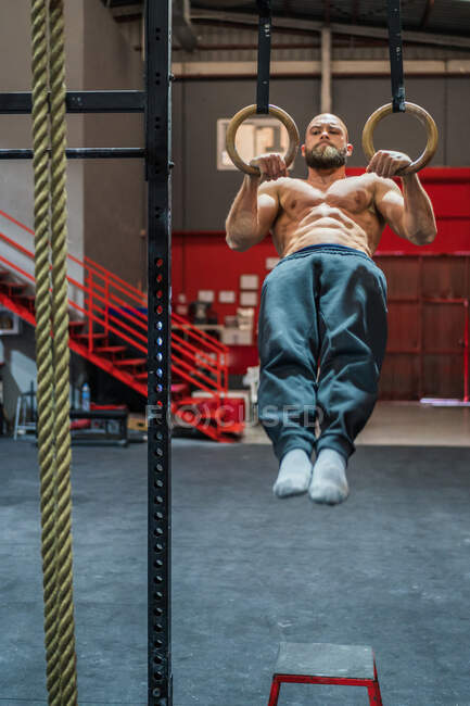 Full length strong shirtless man standing doing exercise on gymnastic rings during intense workout in modern gym — Stock Photo