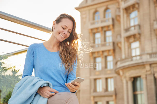 D'en bas de dame adulte souriante en vêtements décontractés debout près de la clôture et du vieil immeuble tout en surfant sur le téléphone dans le quartier de la ville en plein jour — Photo de stock