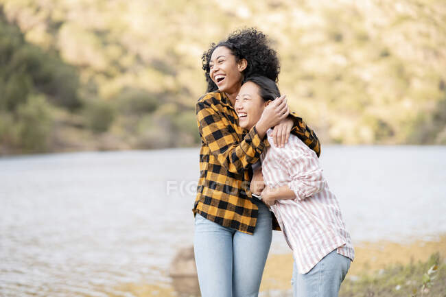 Zufriedenes LBGT-Paar von multirassischen Weibchen, die sich umarmen, während sie mit Hund am See im Wald spazieren gehen und wegschauen — Stockfoto