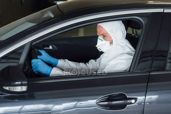Vue latérale d'un biologiste sérieux homme utilisant un masque de protection, des gants et des vêtements tout en conduisant une voiture pendant la période de quarantaine — Photo de stock