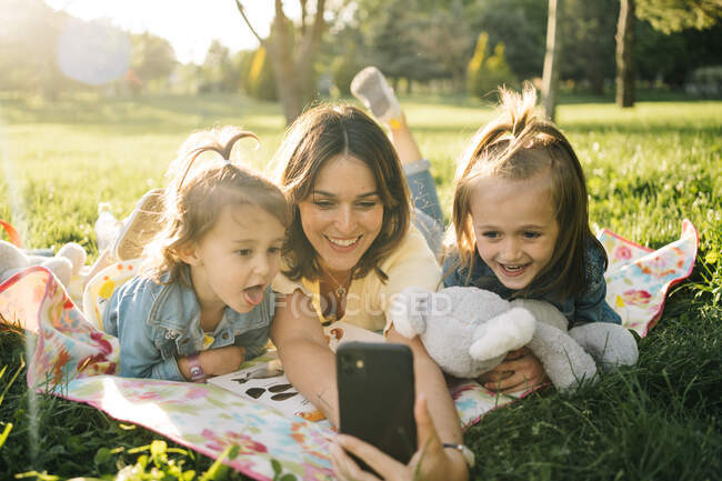 Joyeux jeune femme et mignonnes petites filles allongées sur une couverture et prenant selfie sur smartphone tout en s'amusant ensemble sur la prairie verte dans le parc d'été — Photo de stock