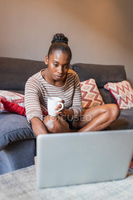 Молода чорна жінка з чашкою гарячого напою, що переглядає Інтернет на нетбуці, сидячи на дивані в кімнаті — стокове фото