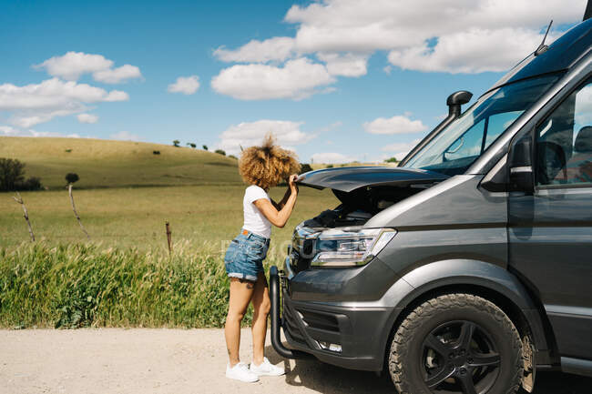 Side view of unrecognizable traveling female looking under opened hood of camper van while having problems during trip through summer nature — Stock Photo