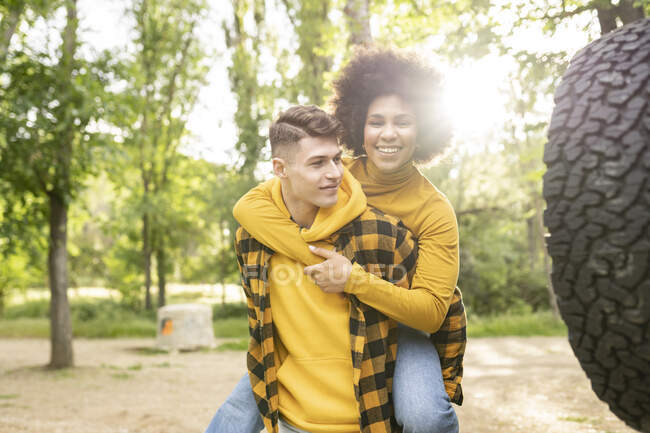 Young man giving piggyback ride to optimistic black girlfriend casual clothes while standing near car on sunny day in countryside — Stock Photo