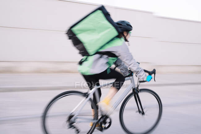 Side view of anonymous female courier with thermal bag riding bike on street road while delivering food in city, motion blur — Stock Photo