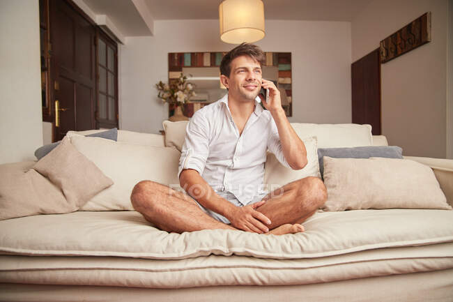 Hombre adulto con ropa casual sentado en el sofá con almohadas mientras conversa en el teléfono inteligente en la sala de estar de luz mientras mira hacia otro lado - foto de stock