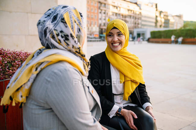 Encantadas amigas muselinas en hijabs sentadas en el banco y mirándose mientras pasan el fin de semana en la ciudad - foto de stock