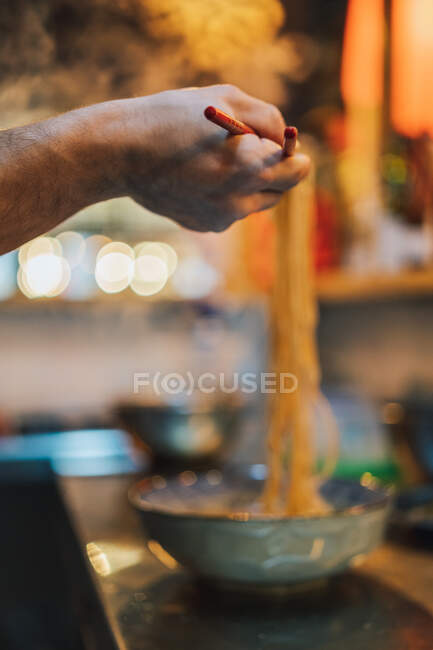 Mão de corte de chef sem rosto com pauzinhos de madeira segurando macarrão acima da tigela no fundo borrado no bar ramen — Fotografia de Stock