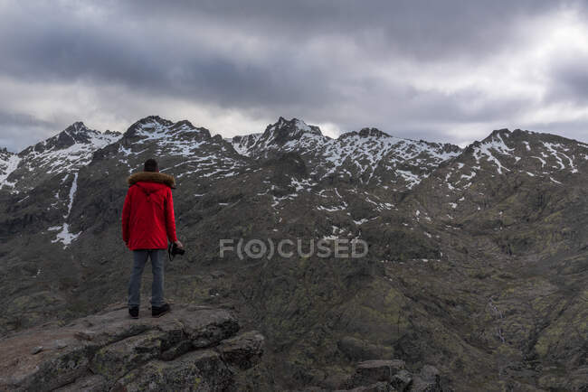 Vista trasera de un hombre irreconocible en ropa de abrigo de pie sobre piedra y mirando la nevada cresta de la Sierra de Gredos en la noche nublada en Ávila, España - foto de stock