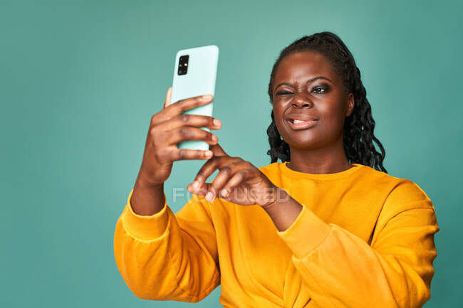Happy plump afro-americano fêmea em camisola amarela piscando e tirando selfie no smartphone contra a parede azul em estúdio — Fotografia de Stock