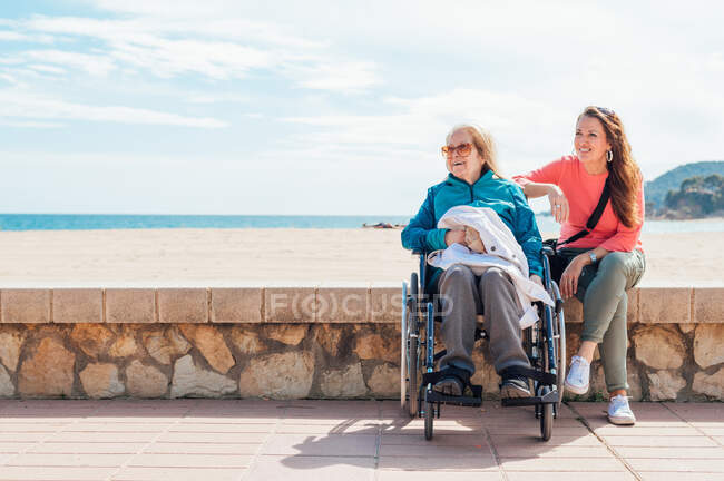 Веселая взрослая дочь с пожилой матерью в инвалидной коляске сидит каменный забор вдоль набережной рядом с морем летом глядя в сторону — стоковое фото