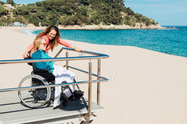 Seitenansicht einer erwachsenen Frau mit älterer Mutter im Rollstuhl mit Blick auf das Meer an sonnigen Tagen — Stockfoto