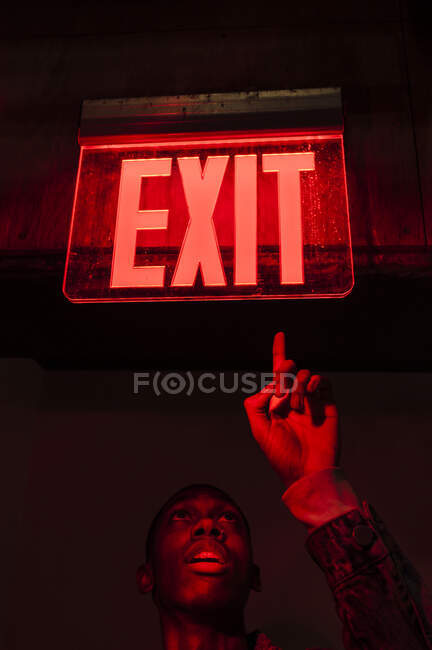 З - під врожаю афроамериканець дивиться вгору на ілюміновану табличку Вихід над головою у червоному темному світлі. — стокове фото