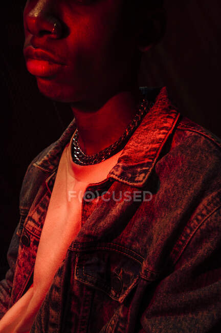Crop calmo homem afro-americano elegante em jaqueta de jeans sob luz vermelha néon na sombra no fundo preto — Fotografia de Stock