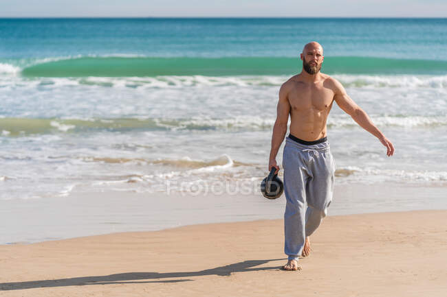Hombre deportivo sin camisa llevando kettlebell caminando por la costa arenosa con olas oceánicas en el fondo mirando hacia otro lado - foto de stock