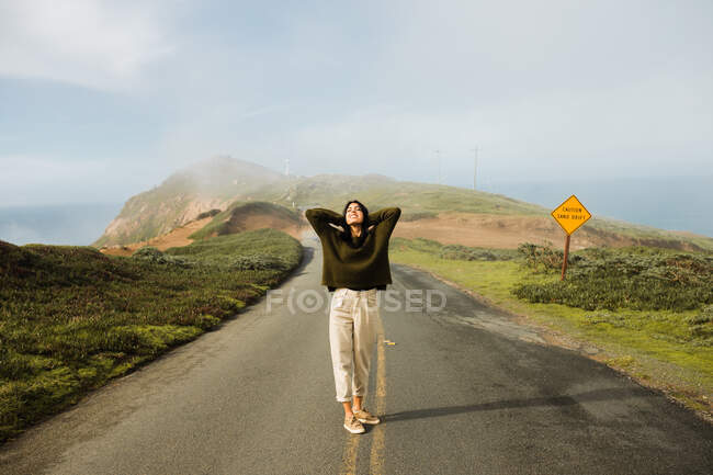 Full length of cheerful woman holding hands behind head in enjoyment of sunlight standing on empty route of coastal cliff in California — Stock Photo
