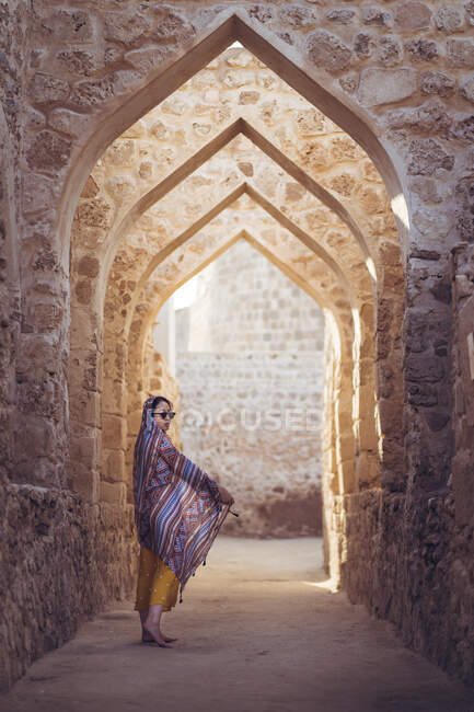 Mujer vestida con ropa tradicional oriental caminando a través de una larga sala arqueada con columnas de piedra del famoso Qal 'at al-Bahrain - foto de stock