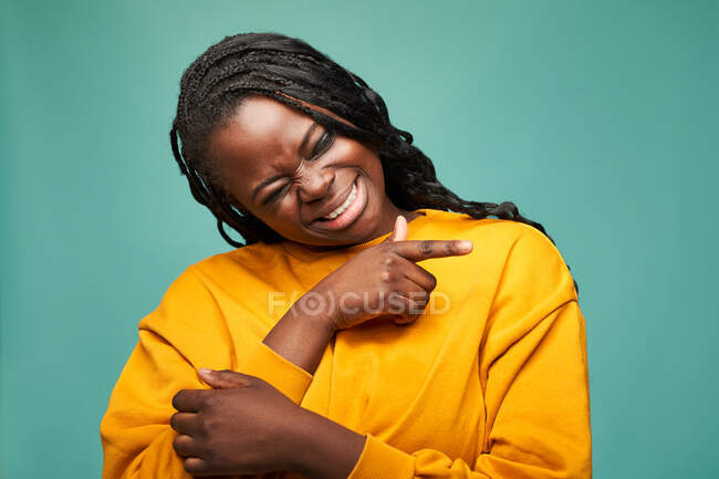 Glad African American female in yellow clothes with eyes closed pointing finger away against blue background — Stock Photo