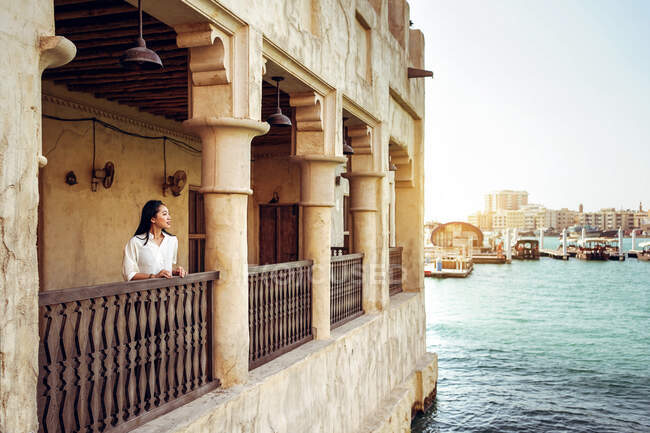 Junge Touristinnen stehen auf der Terrasse eines alten Steingebäudes direkt am Wasser und genießen die Stadtlandschaft, während sie das historische Viertel Al Fahidi in Dubai besuchen. — Stockfoto