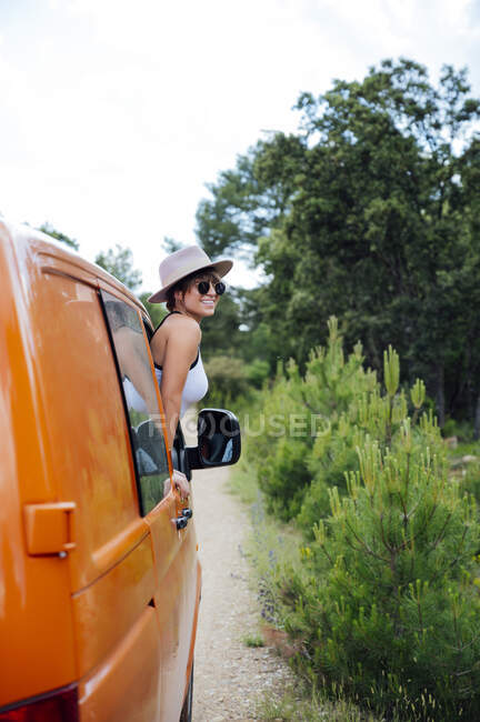 Веселая женщина-путешественница, выглядывающая из окна фургона и показывающая два пальца жестом, наслаждаясь поездкой летом — стоковое фото