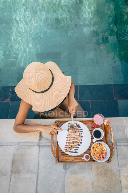 Von oben eine anonyme Touristin mit Strohhut, die im Pool sitzt und köstlichen Crêpe mit Schokoladensoße schneidet — Stockfoto
