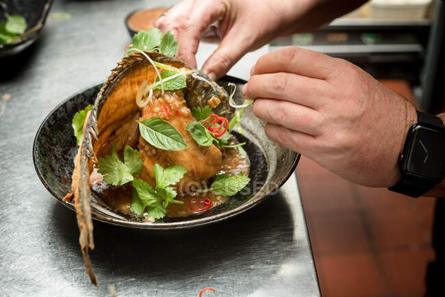 Crop anonymous male chef garnishing deep fried fish with green herbs on plate in restaurant — Stock Photo